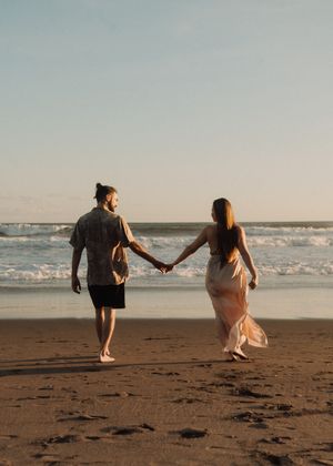 A couple holding hands while walking along a sandy beach at sunset with ocean waves in the background.