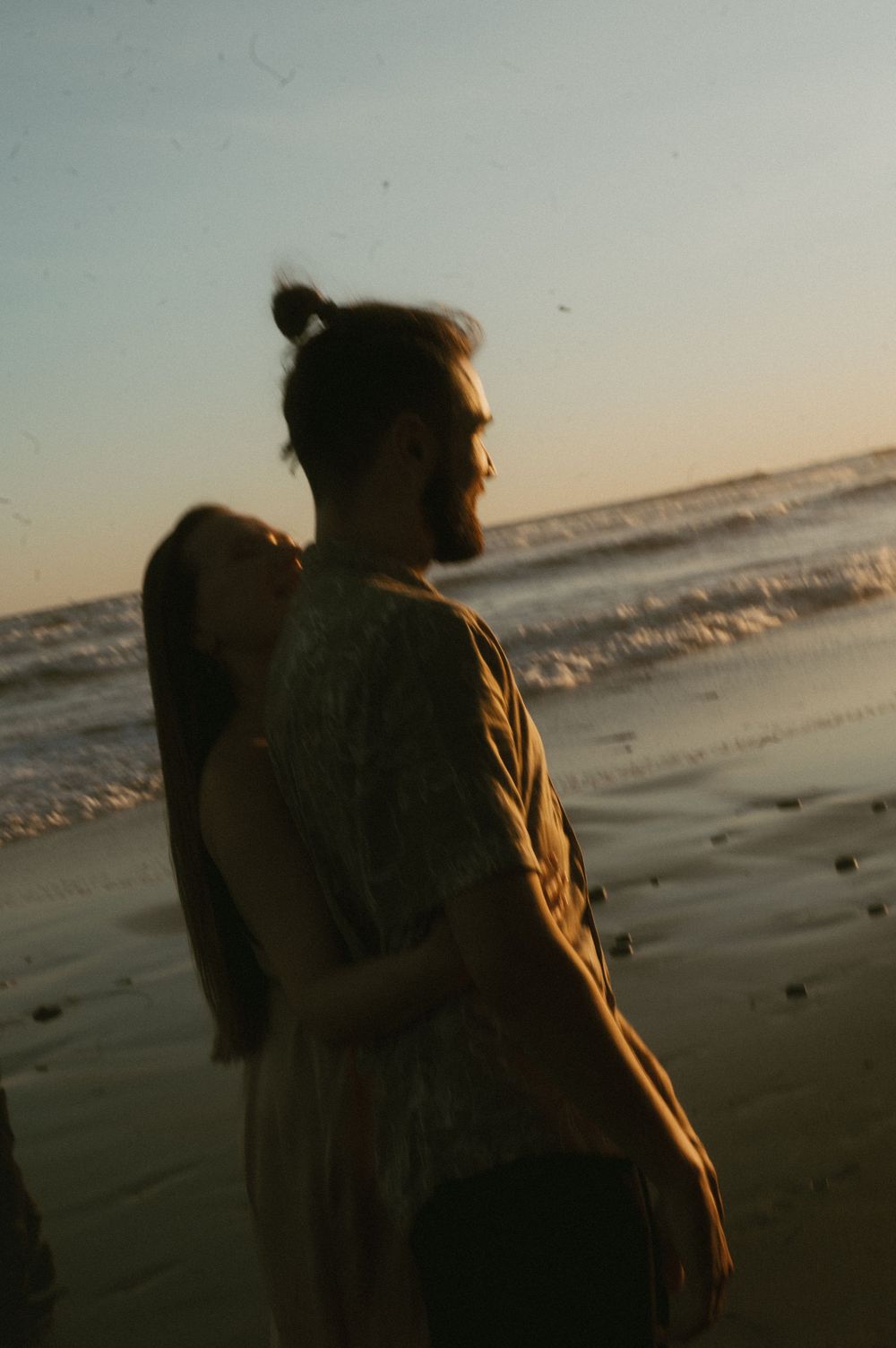Silhouetted figure in a brown jacket stands on a beach at sunset with ocean waves in the background.