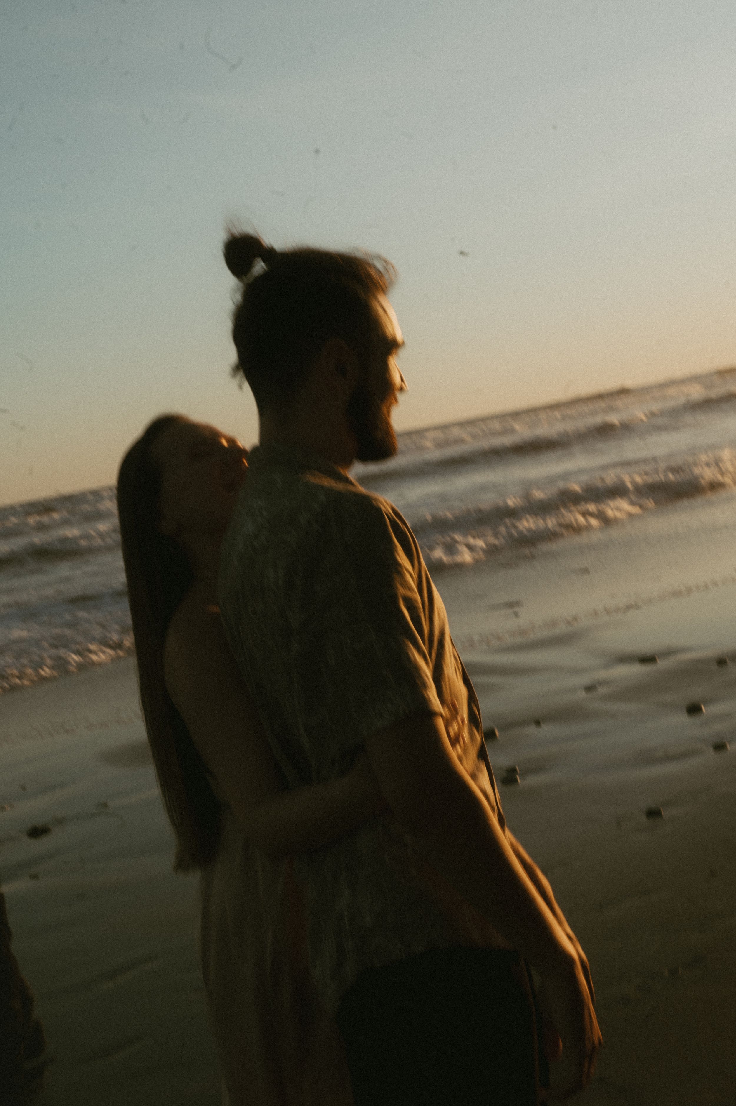Silhouetted figure in a brown jacket stands on a beach at sunset with ocean waves in the background.