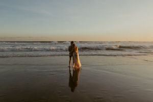 A solitary figure stands on a reflective beach at sunset as waves roll in from the ocean.
