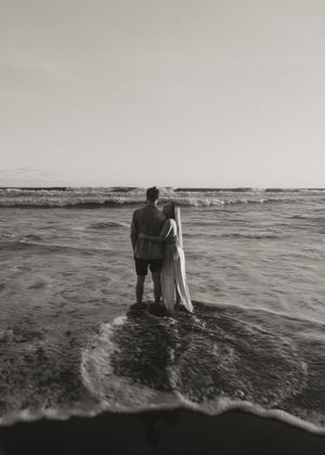 A romantic couple stands together in shallow ocean waves at sunset in black and white.