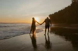 Silhouettes of two people walking hand in hand along a beach at sunset with waves crashing in the background.