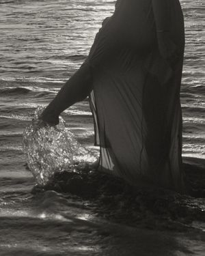A figure in a flowing black dress stands in shallow ocean water creating splashes in dramatic black and white photography.