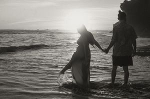 Two silhouetted figures hold hands while standing in shallow ocean waves at sunset.