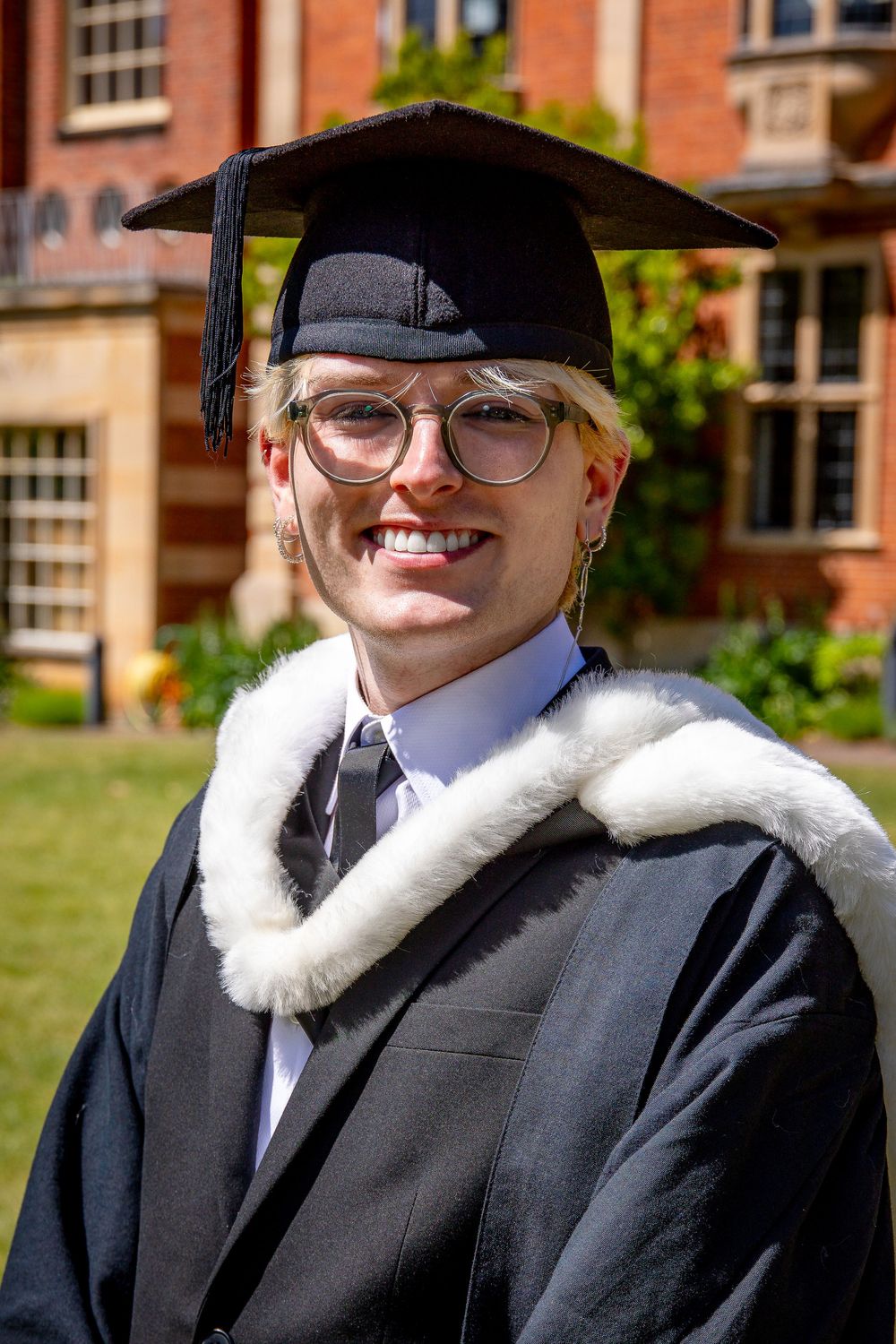 Graduate in academic regalia with white stole and mortarboard smiles on sunny campus day.
