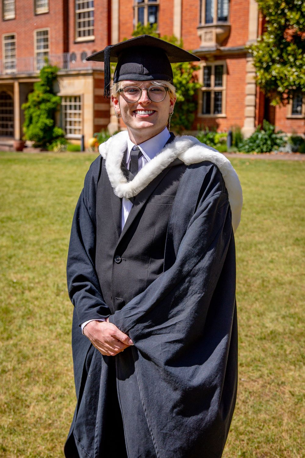 Graduate in traditional black academic dress stands on sunny lawn near historic building.