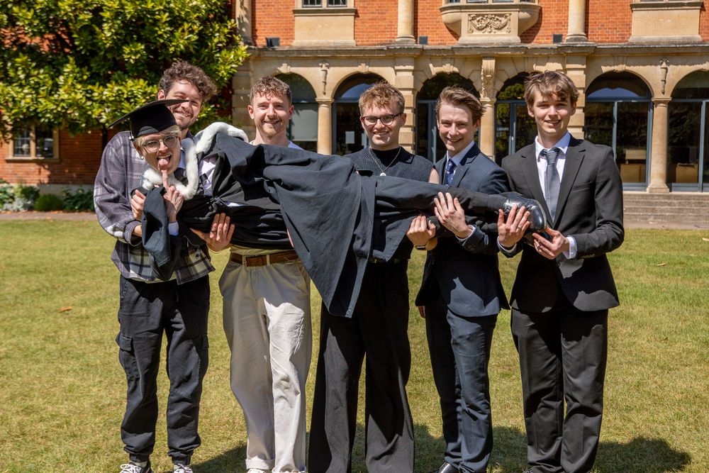 Group of graduates in formal attire pose together on lawn in front of red brick building.