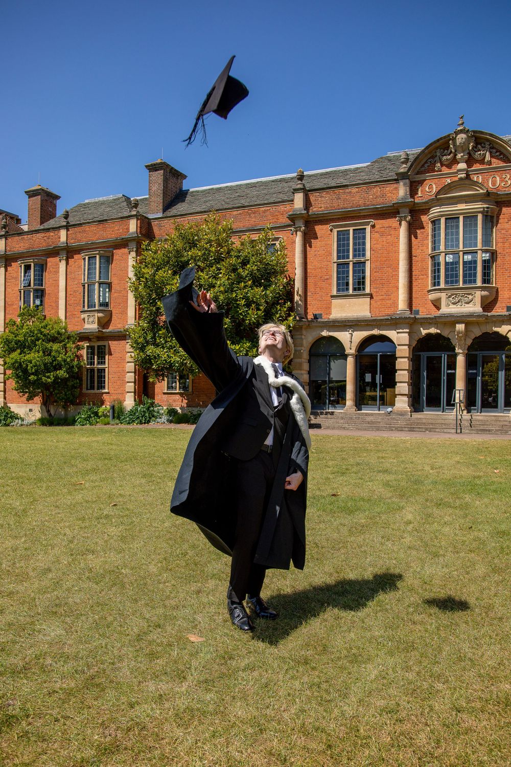 Graduate in black academic regalia throws cap in celebration on sunny campus lawn.