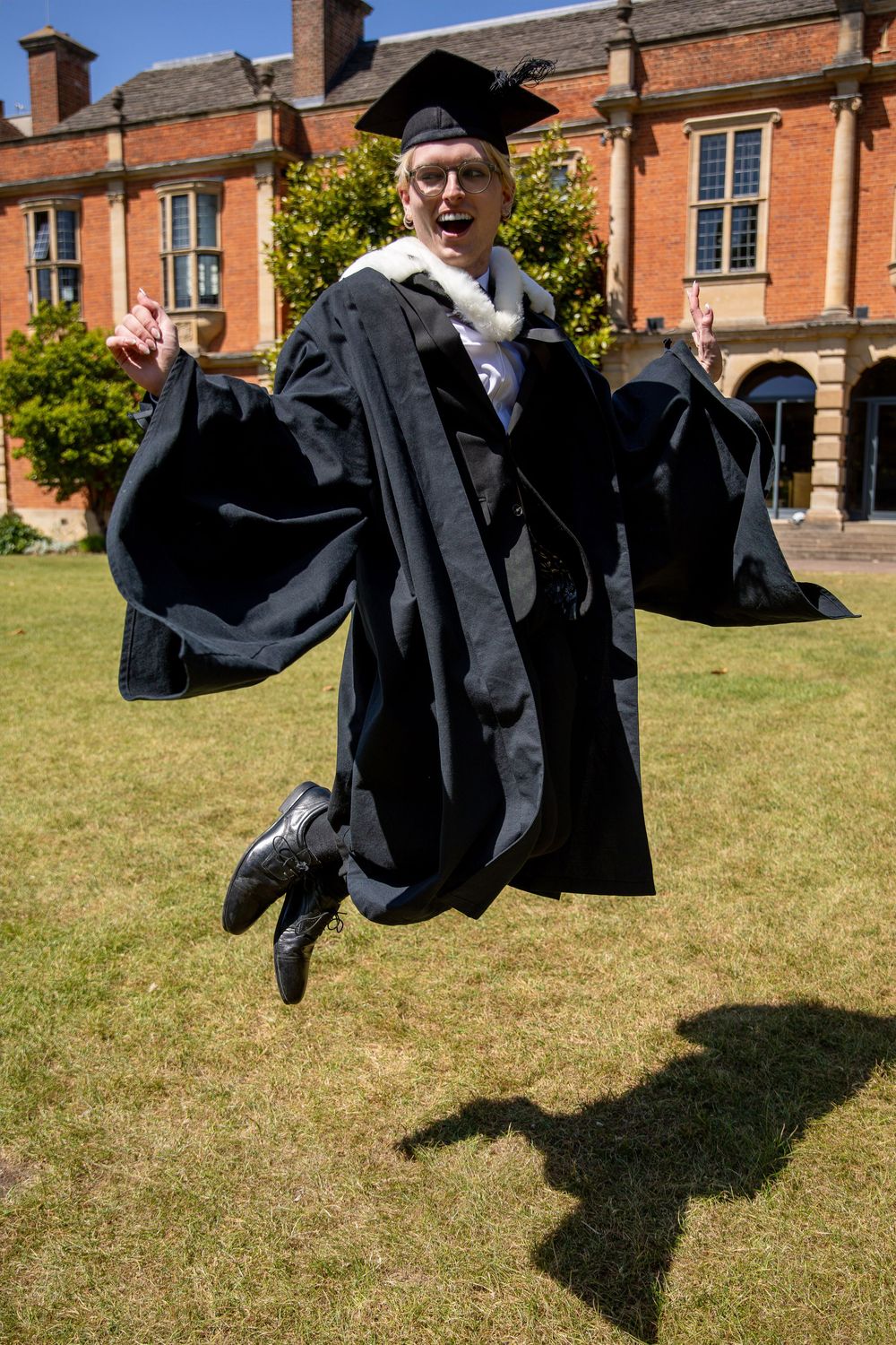 Graduate leaps joyfully in academic regalia on sunny campus lawn.