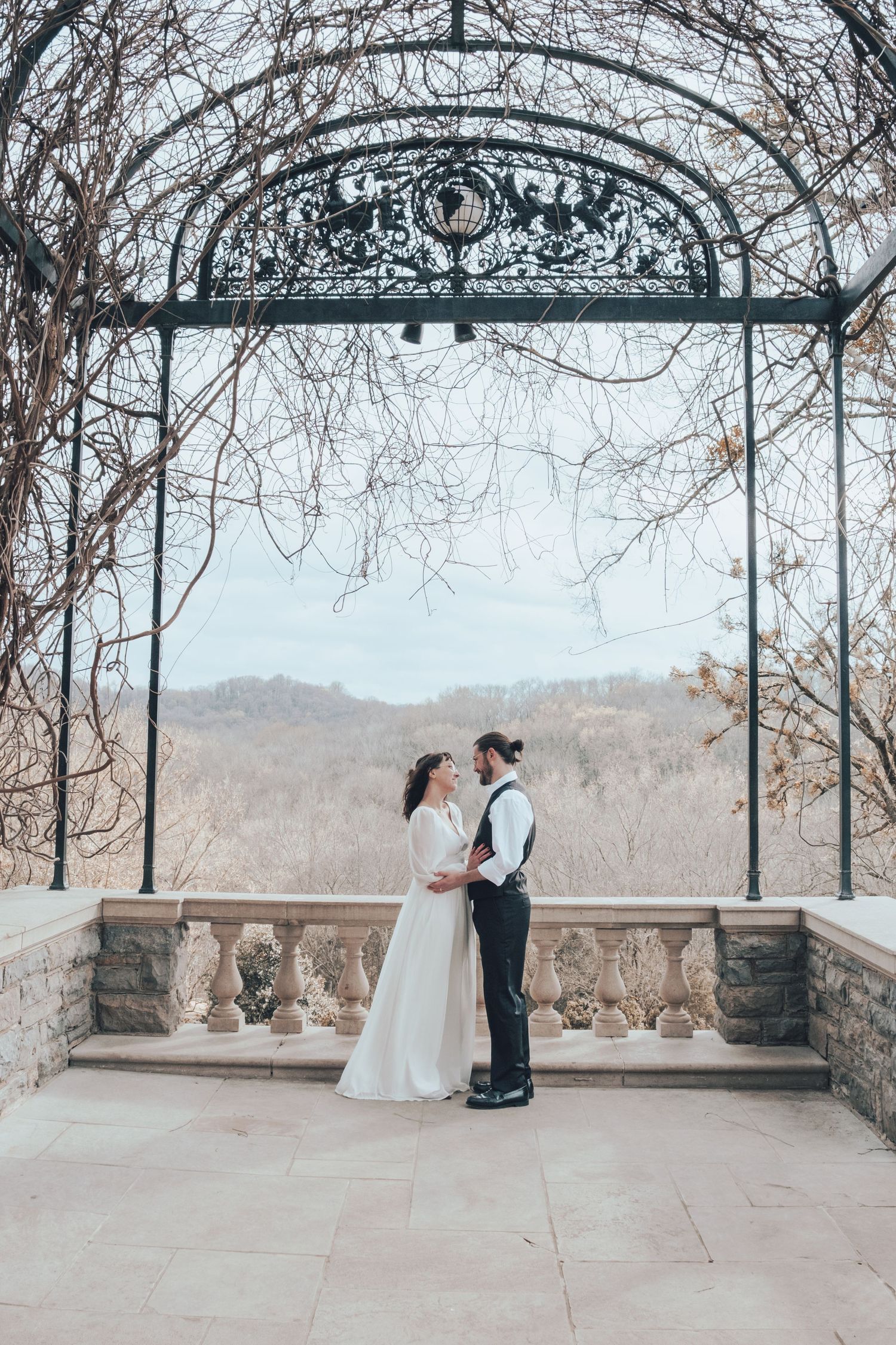 Elegant wedding couple standing under an ornate metal archway overlooking a scenic mountain vista.