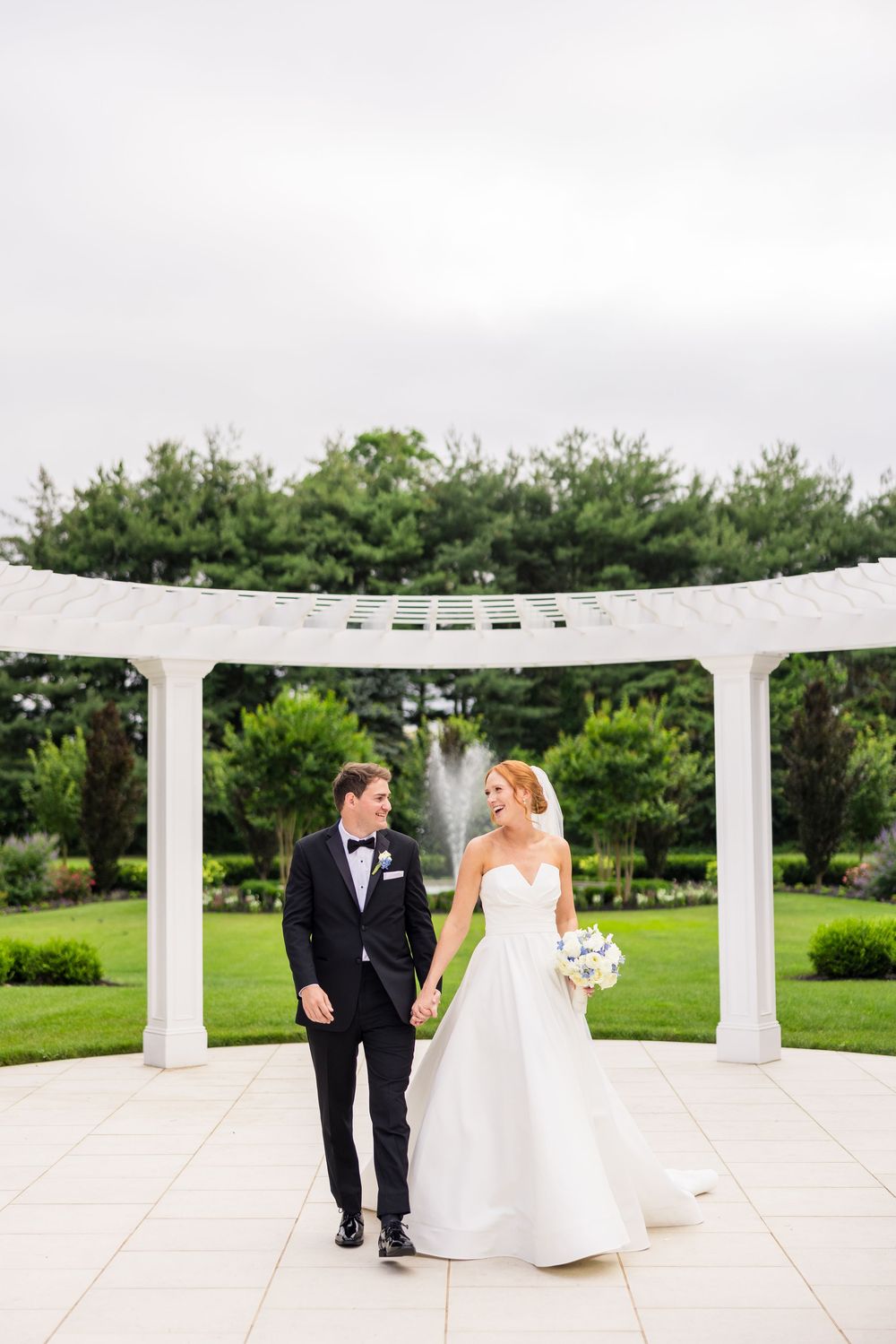 A couple in formal wedding attire stroll together on a white paved terrace with elegant pillars and green landscaping.