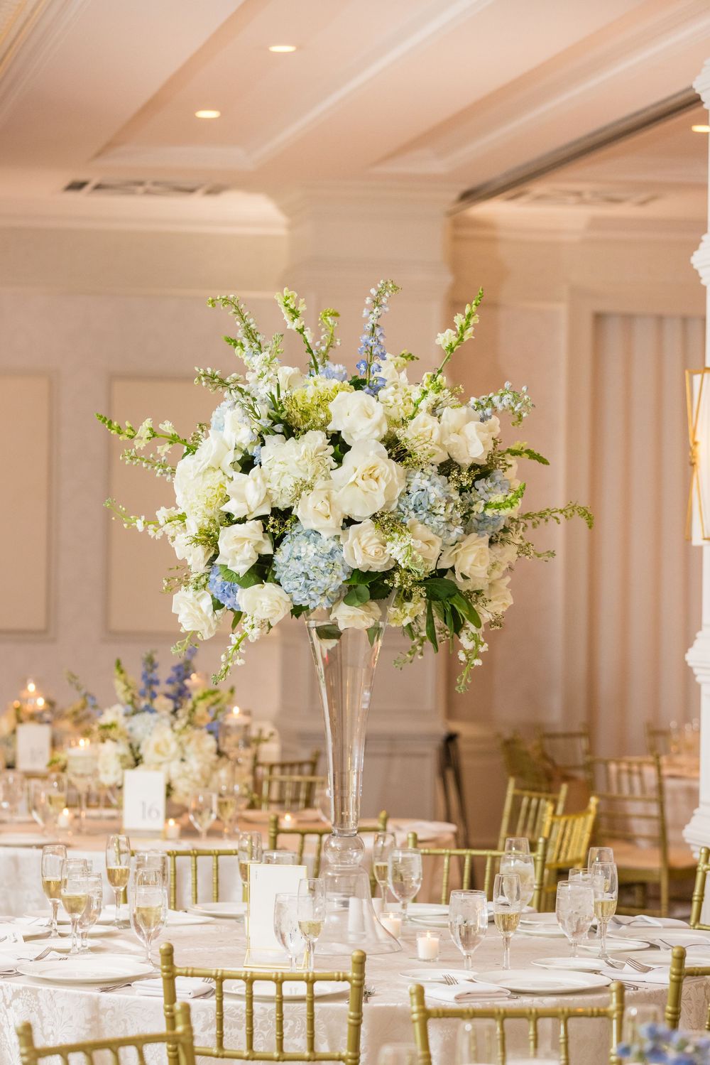 Elegant tall floral centerpiece with white and light blue flowers in crystal vase on decorated wedding reception table.