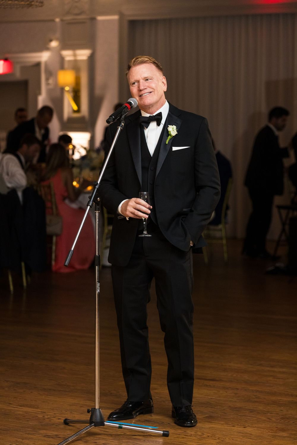 A person wearing a black tuxedo holds a drink while standing on a wooden dance floor at a formal event.