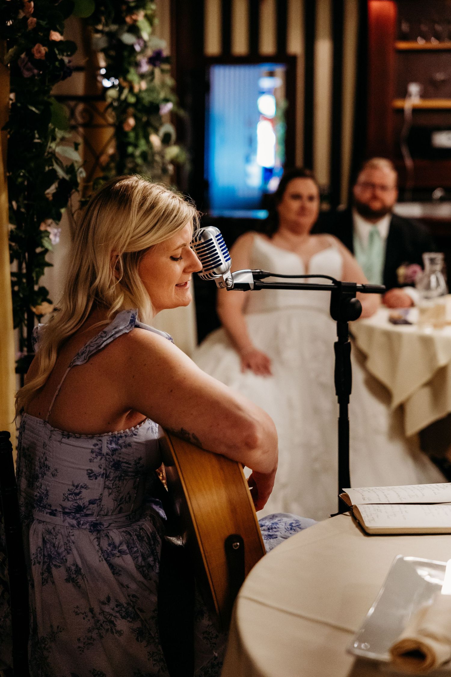 A musician in a floral dress performs with an acoustic guitar at an intimate wedding reception dinner.