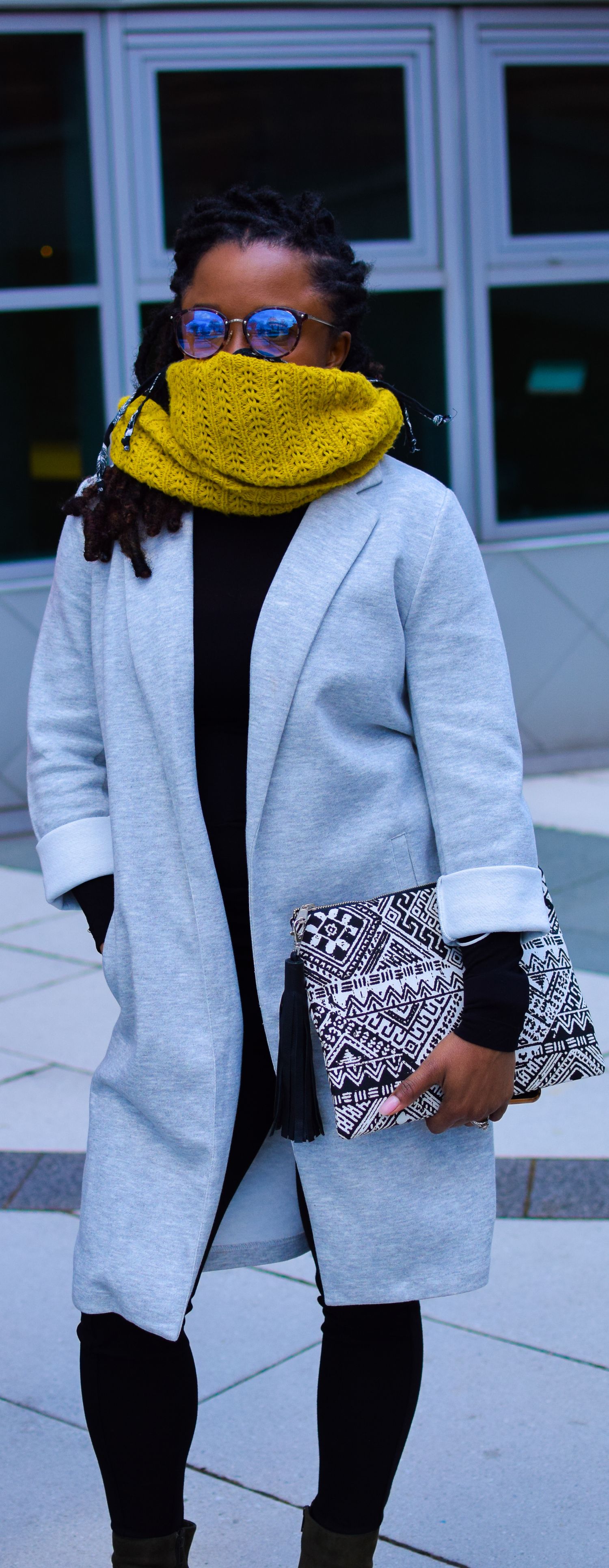 Person wearing gray coat, yellow knit scarf, blue sunglasses and carrying black-white patterned clutch stands outdoors.