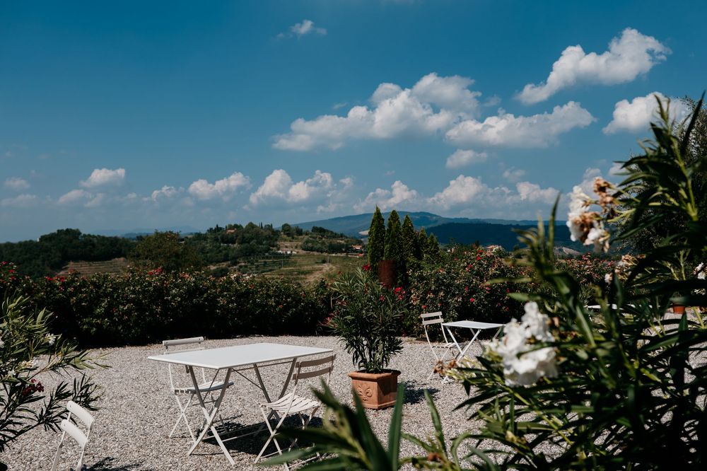 Outdoor patio with white bistro tables overlooking Tuscan hills and mountains against a bright blue sky with fluffy clouds.