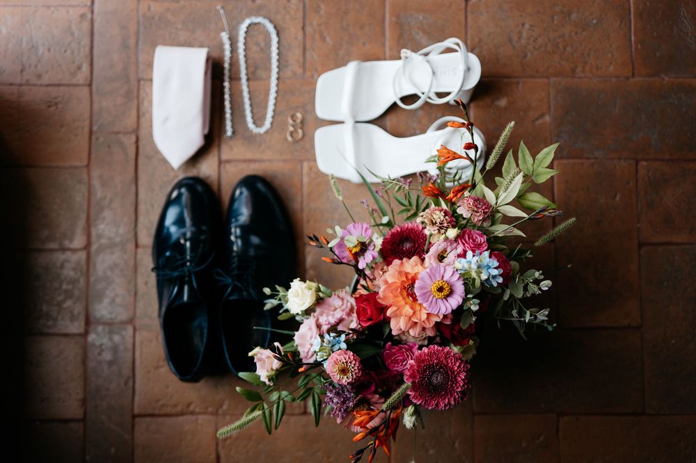 Wedding essentials arrangement showing black dress shoes, white heels, floral bouquet with pink and red flowers on terracotta tiles.