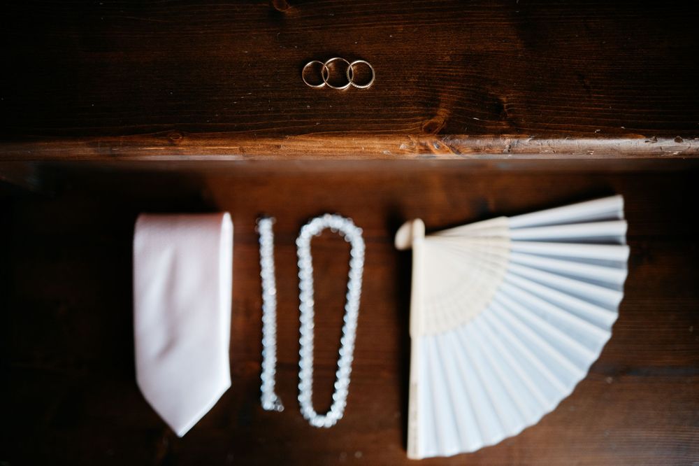 Wedding accessories including white tie, pearl necklace, and folding fan arranged on dark wooden surface with rings above.