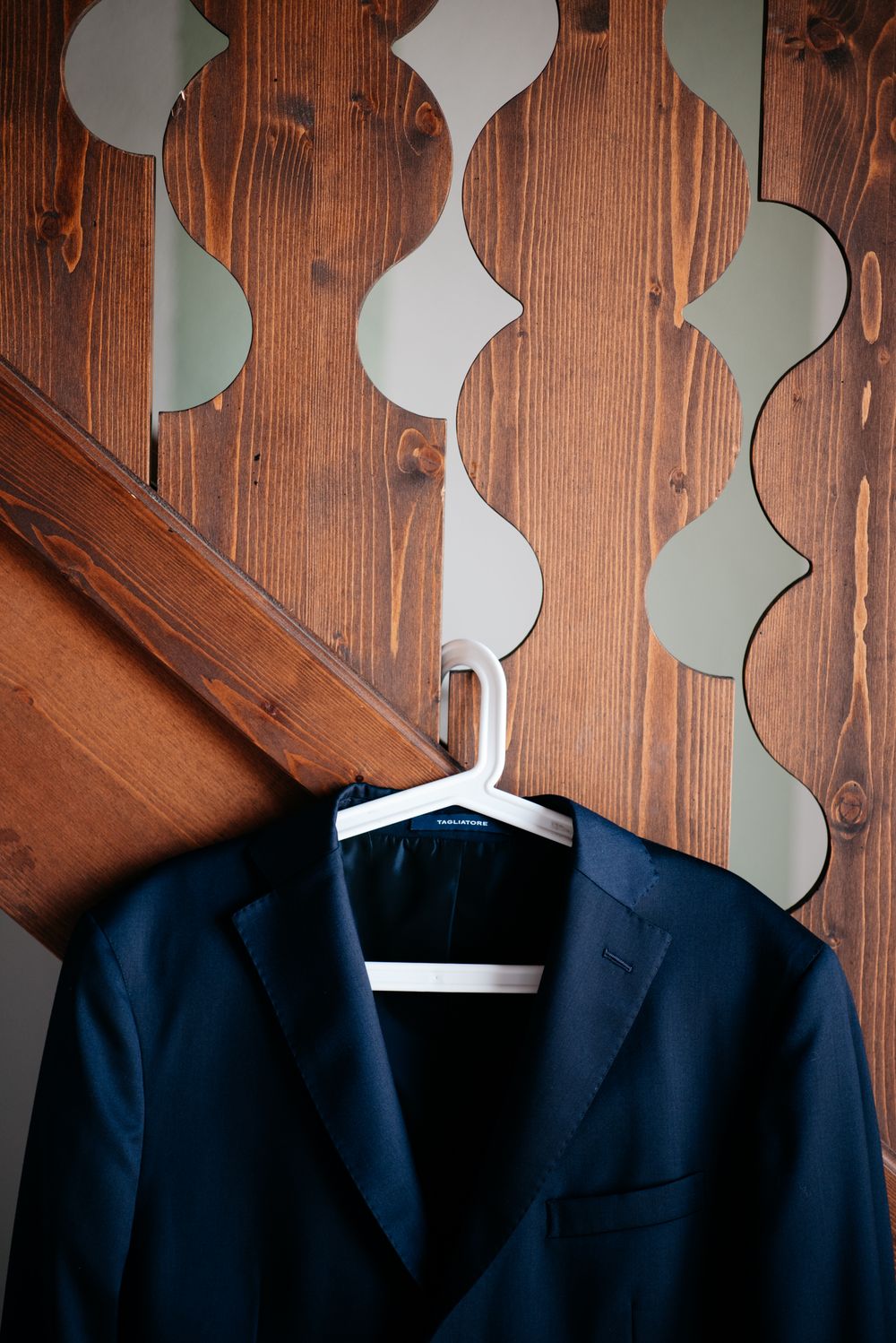 Navy blue formal suit jacket hanging on white hanger against wooden wall with decorative curved patterns.