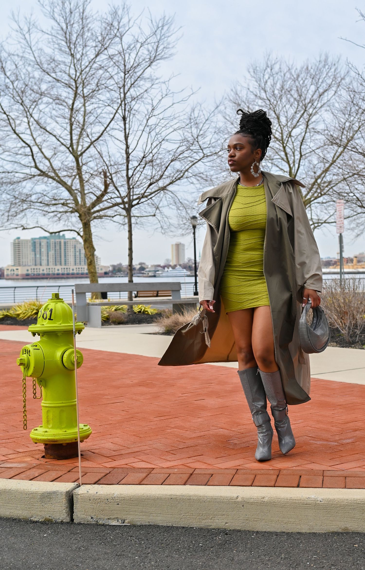 A stylish woman in a yellow dress and trench coat poses confidently next to a bright yellow fire hydrant on a brick sidewalk.