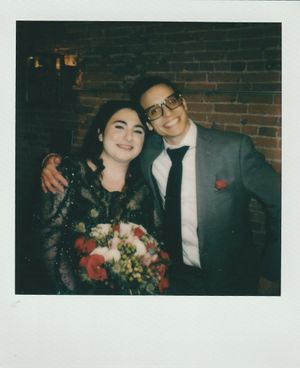A vintage-style polaroid photo of a couple in formal attire with a red floral bouquet against a brick wall background.