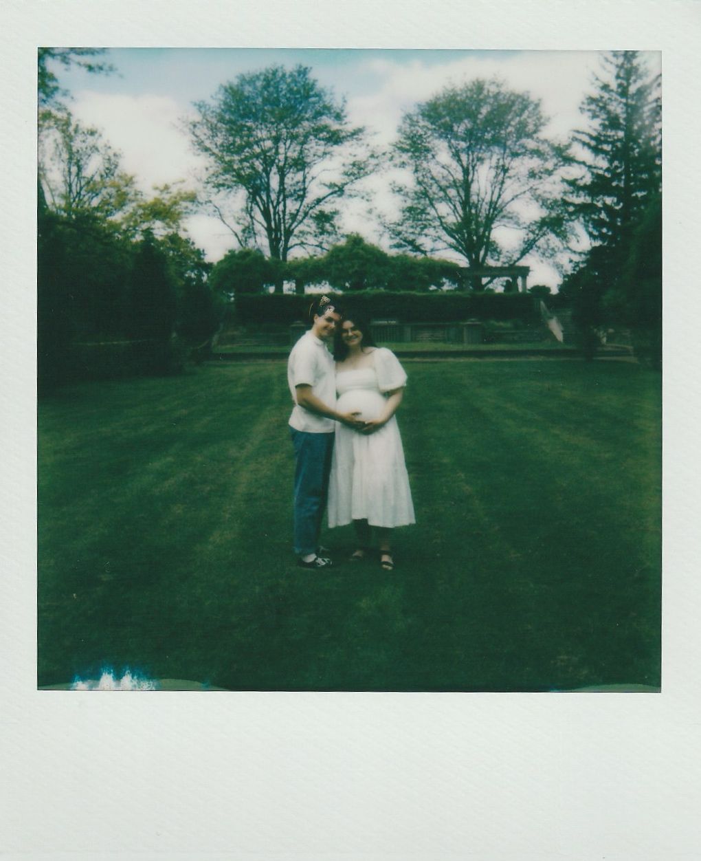 Two people embrace while standing on a manicured lawn with trees in the background on a sunny day.