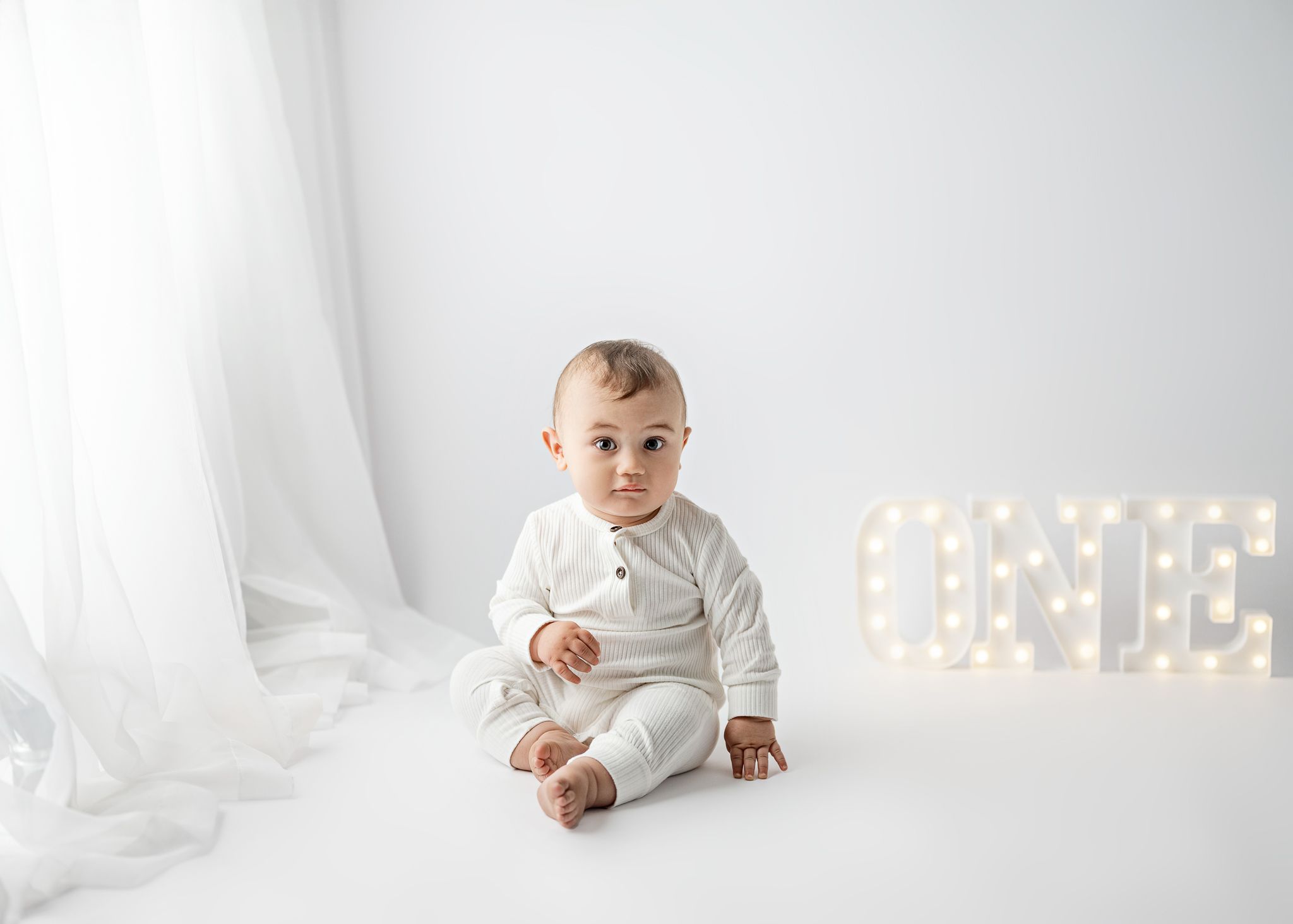 Baby in white outfit sits on white floor with illuminated LOVE marquee sign in background.