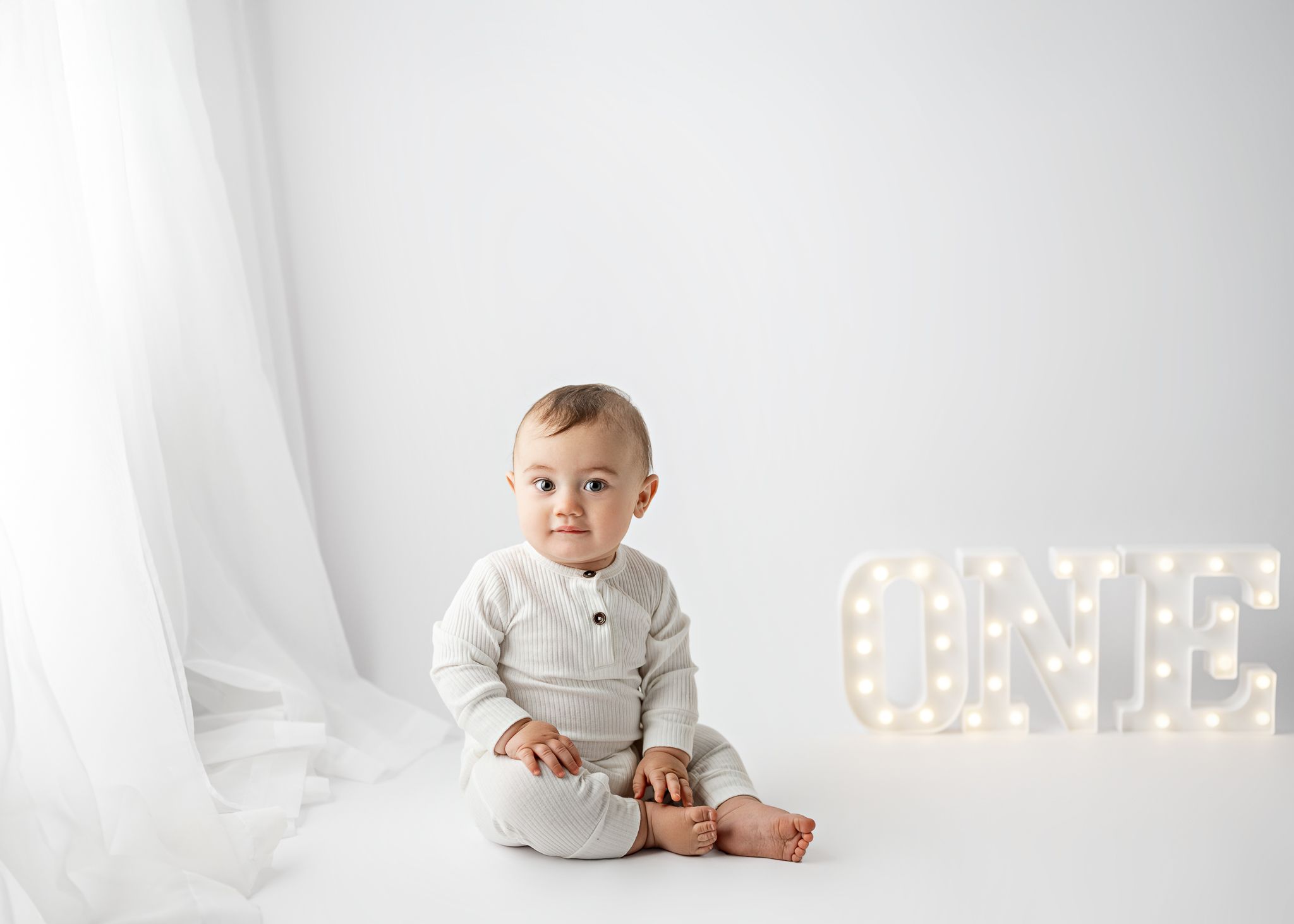 A child in a gray outfit sits on white fabric near illuminated letters in a minimalist studio setting.