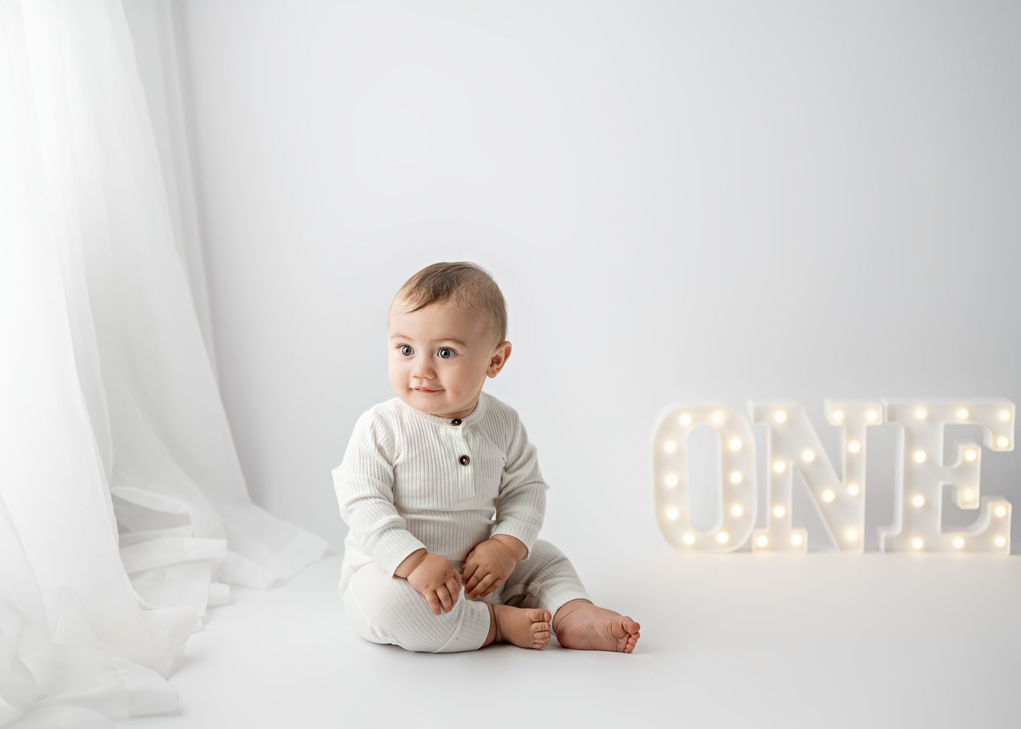 Baby in white ribbed outfit sits on white backdrop with illuminated letters in background.