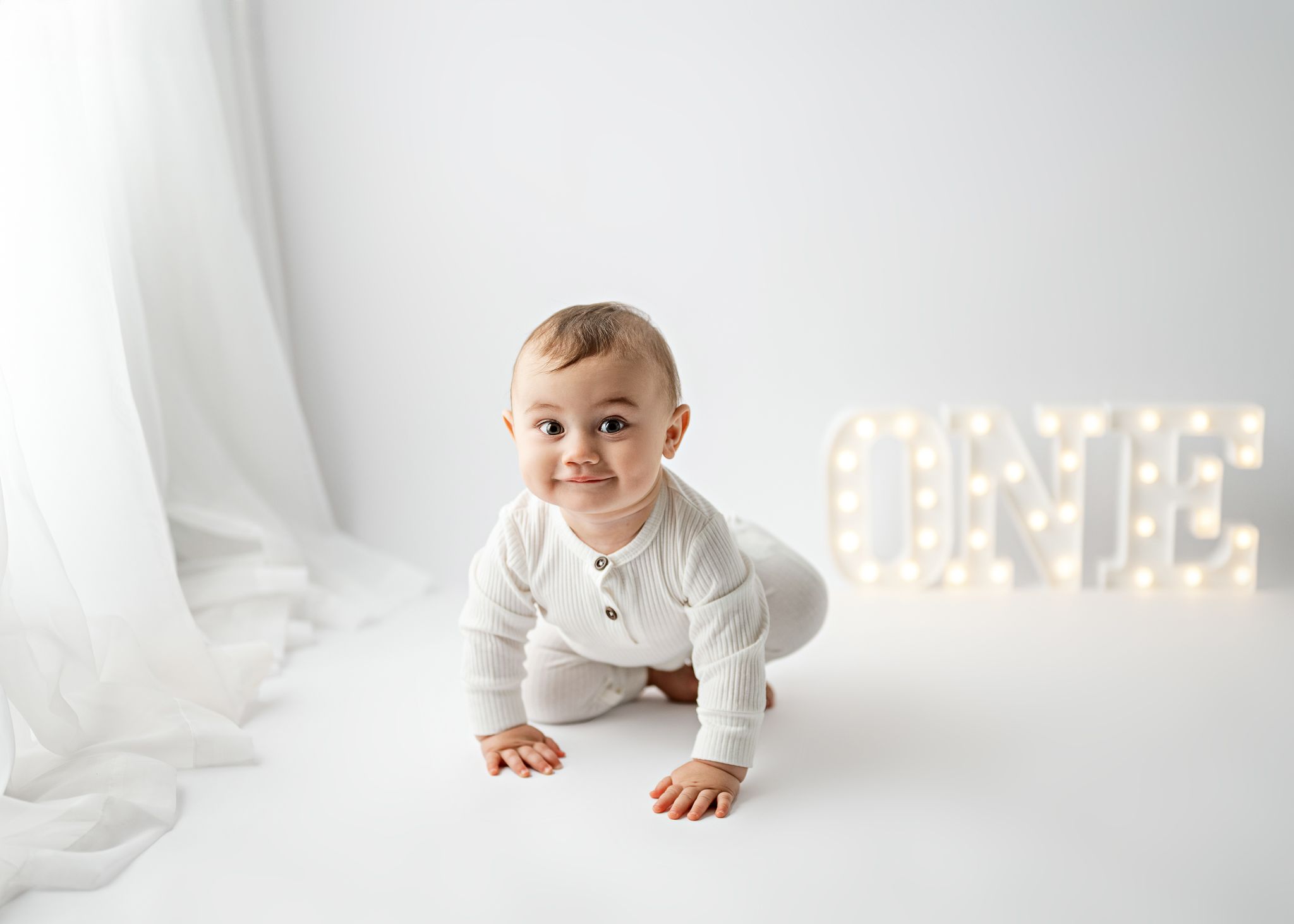 Baby in white romper crawls on white studio setting with glowing marquee letters.