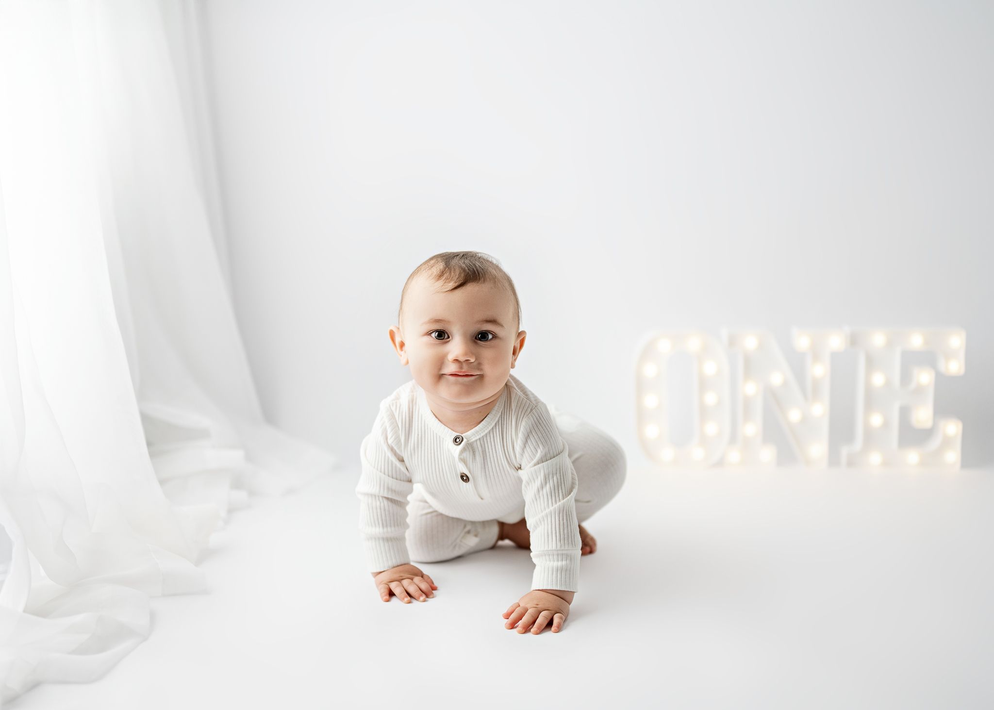 Smiling baby in white outfit crawls on white backdrop with illuminated letter lights in background.