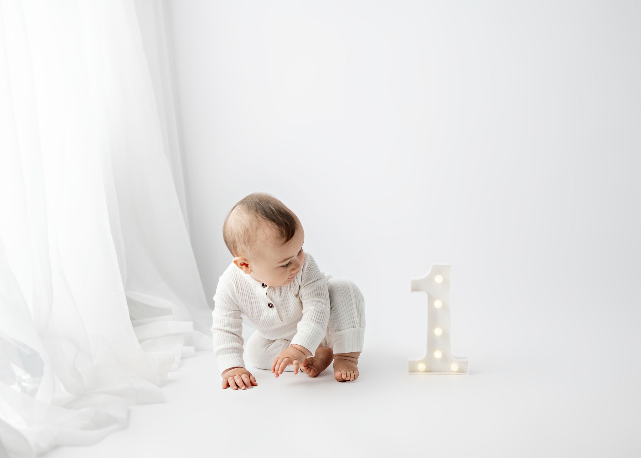 Baby in cream colored outfit crawls on white floor next to number one light decoration.