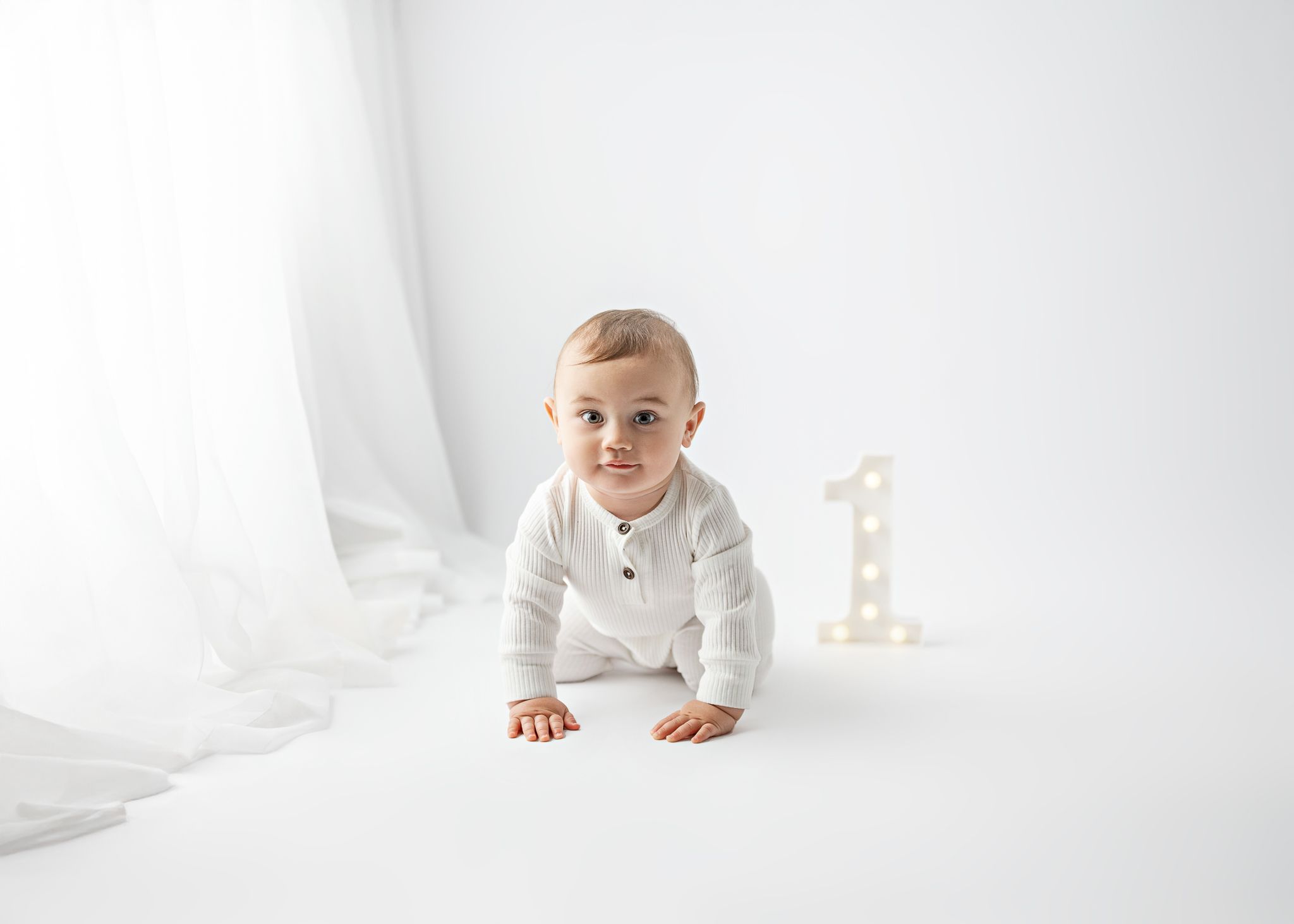 A baby in white clothing crawls playfully on a minimalist bright white backdrop with a number one decoration visible.