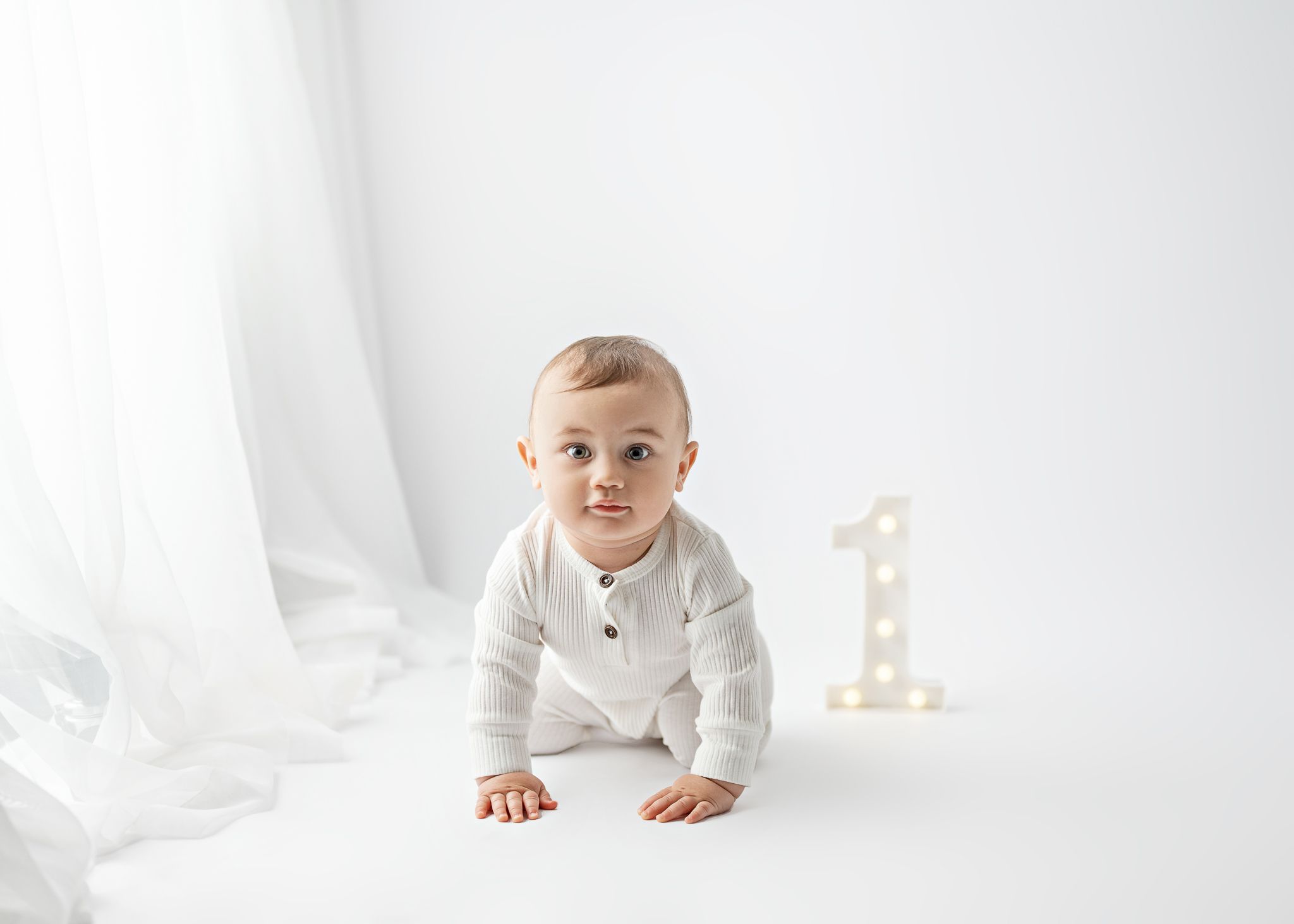 Baby in white sweater crawls on white backdrop next to decorative number one.