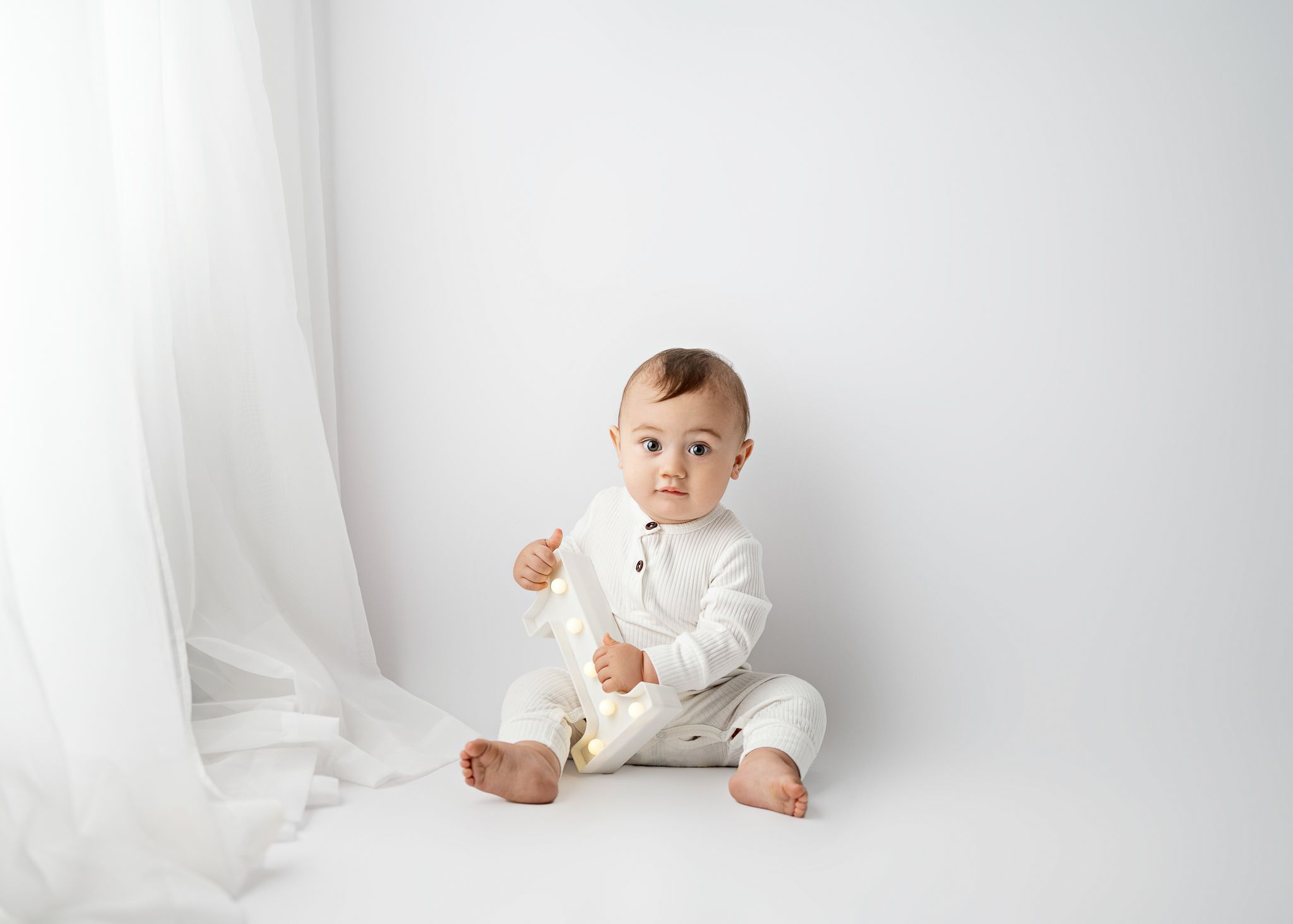 Baby in cream-colored outfit plays while sitting against white background with soft drapery.