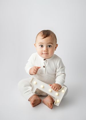 Small baby wearing a cozy white knit outfit sits on plain grey backdrop while holding an illuminated letter block.