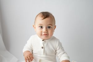 Baby in white knit sweater sits against plain white studio backdrop for portrait.