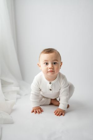 Baby in white knit sweater crawls on white background looking at camera during minimalist studio photoshoot.
