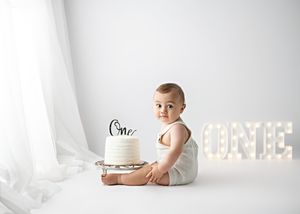 Baby in white outfit sits next to birthday cake with illuminated ONE sign in background for first birthday photoshoot.