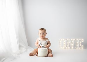 Baby wearing white onesie sits against light backdrop with illuminated 'ONE' sign for first birthday photo session.