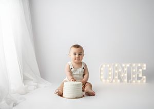 A small child in overalls sits on white fabric beside a cake with lighted 'ONE' marquee letters in the background.