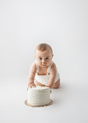 Baby wearing white romper sitting in front of a plain white cake on a white background during baby photo shoot.