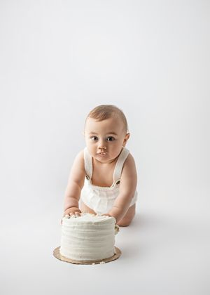A baby in white overalls leans over a simple white frosted cake on a white background.