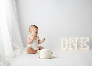 Baby in white onesie sits on white backdrop with birthday cake and illuminated ONE marquee letters.