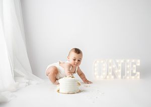 Baby in white onesie smashes first birthday cake while illuminated letters glow in background on white setting.