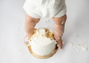 Small hands covered in flour hold a white frosted cake from above on a light backdrop.