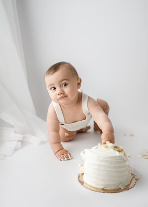 Baby in white romper reaches toward white frosted smash cake during first birthday photo session against white backdrop.