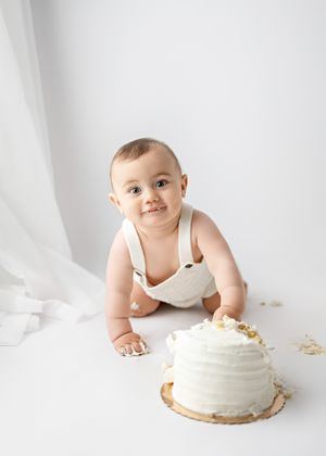 A baby in white clothing crawls beside a beautifully decorated white cake on a light backdrop.