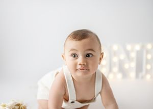 A baby in white overalls crawls on a white surface with glowing decorative lights in the background.