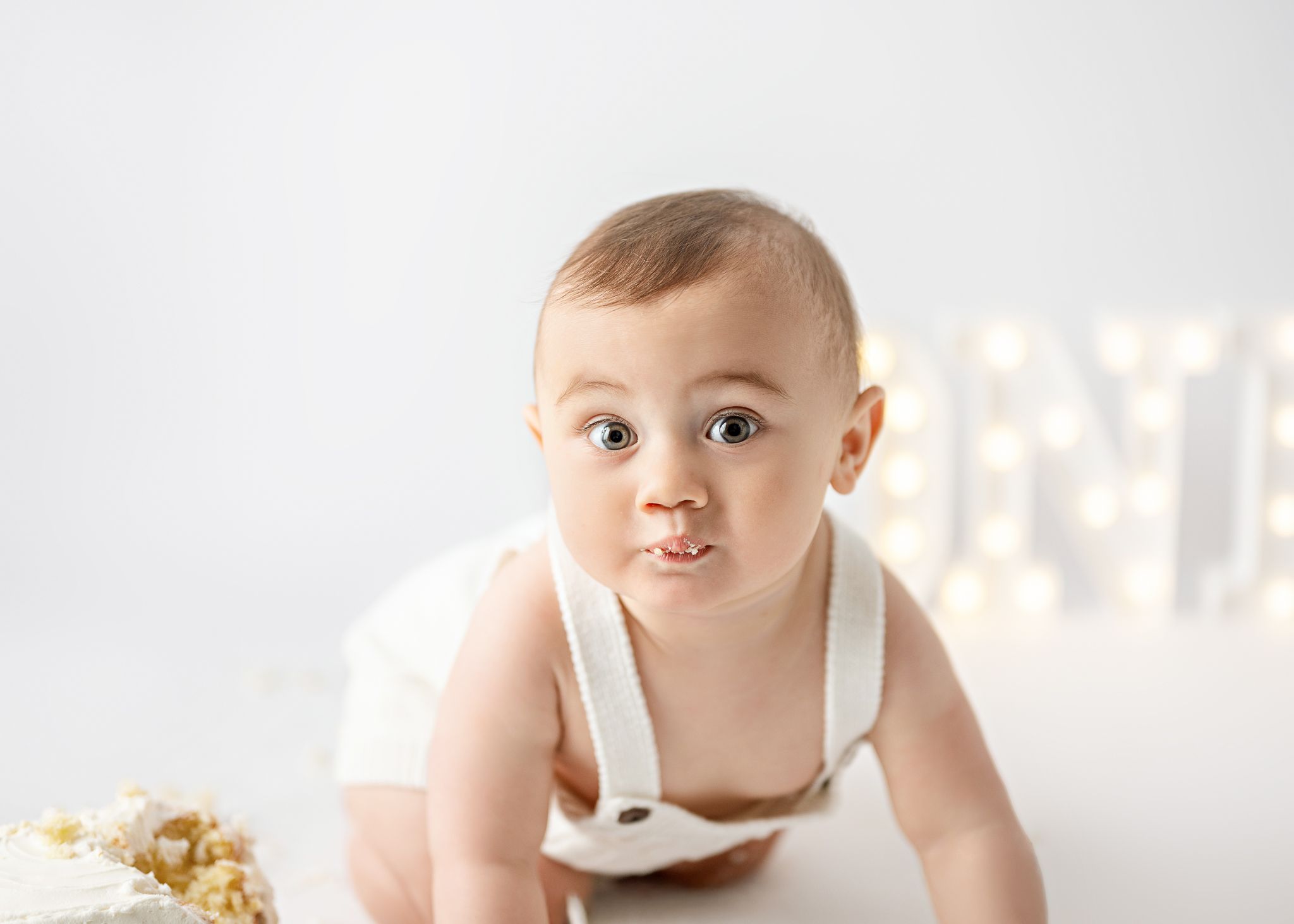 Baby wearing white romper crawls on white backdrop with twinkle lights glowing softly in background.